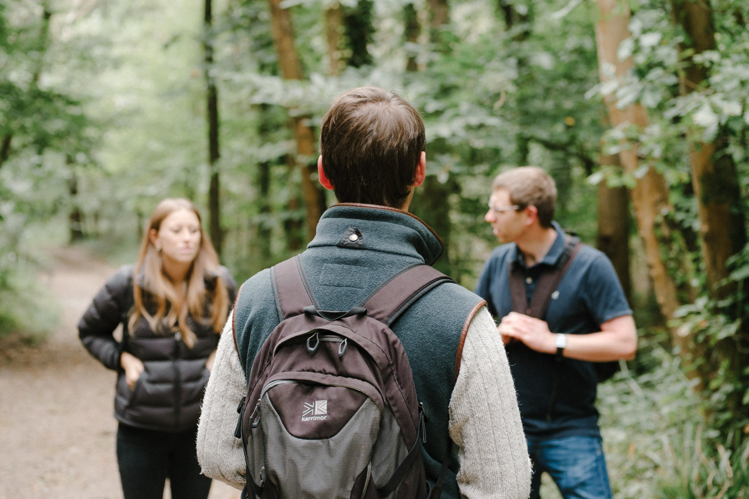 A photo of the back of David's head and backpack. Alice and Adam are in the background but blurred due to the depth of field. All three of them are surrounded by green trees.