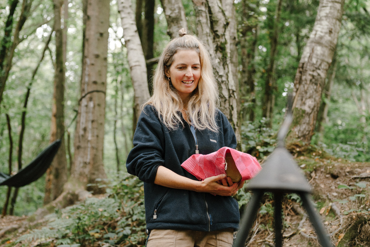 A picture of a blond woman in a grey fleece and tanned trousers. She is holding a mixing bowl with a red tea towel over the top. She is stood in the forest and there's a hammock hanging in the background.