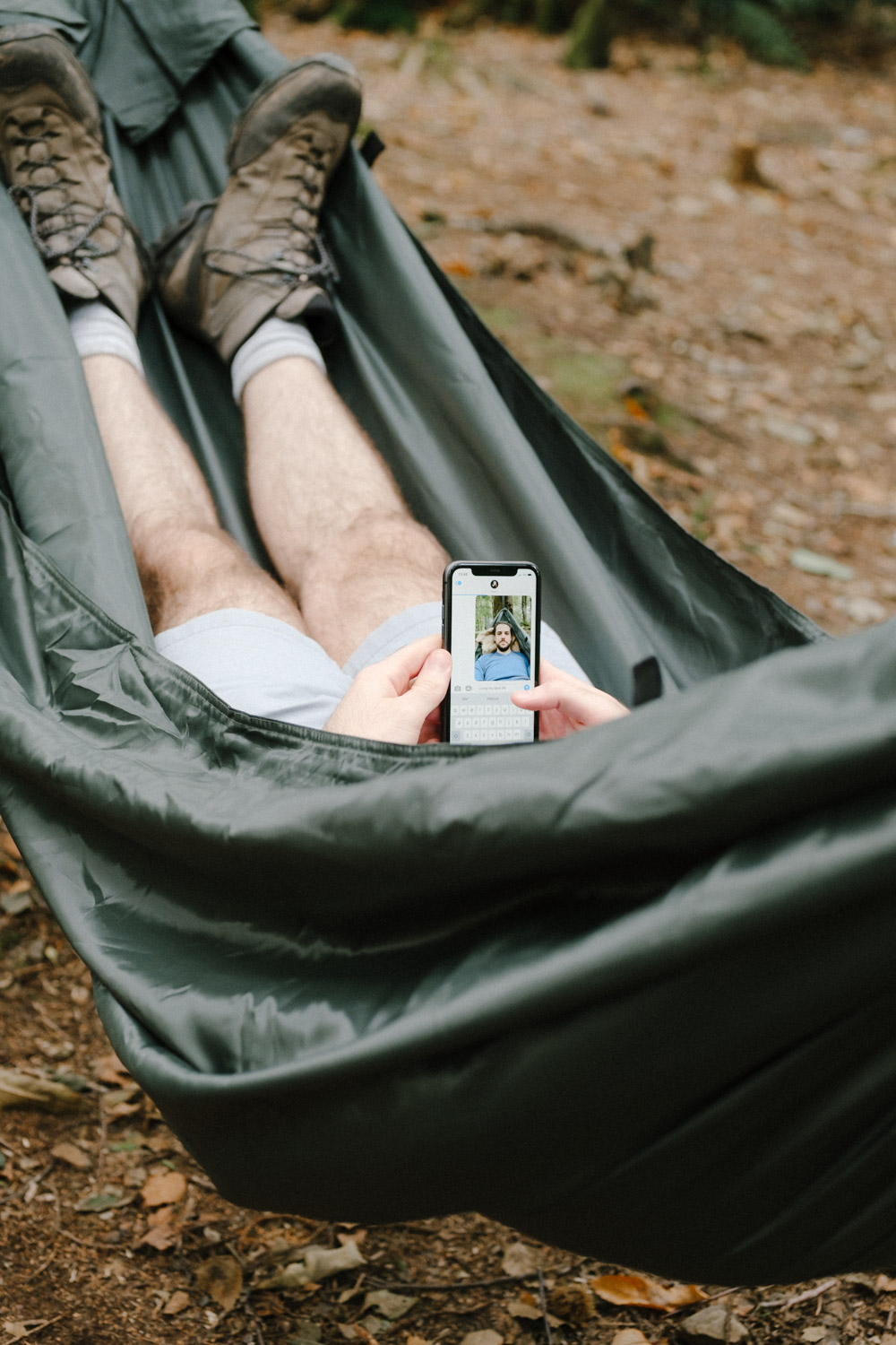 A pair of legs with walking boots on sitting in a hammock. The person is holding their phone up and is taking a selfie.